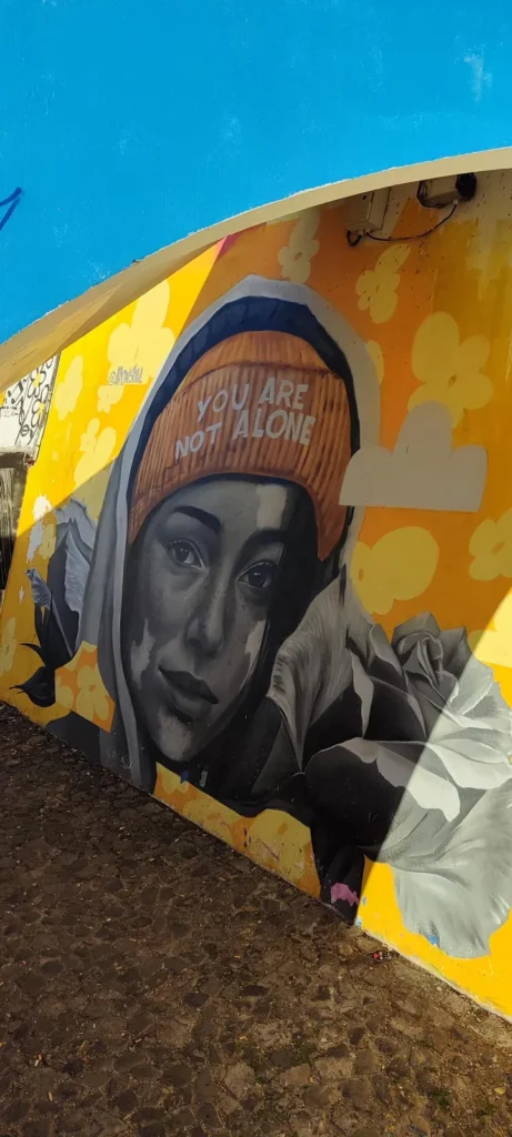 Wandbild einer jungen Frau mit einer orangen Mütze, auf der „You are not alone“ steht.